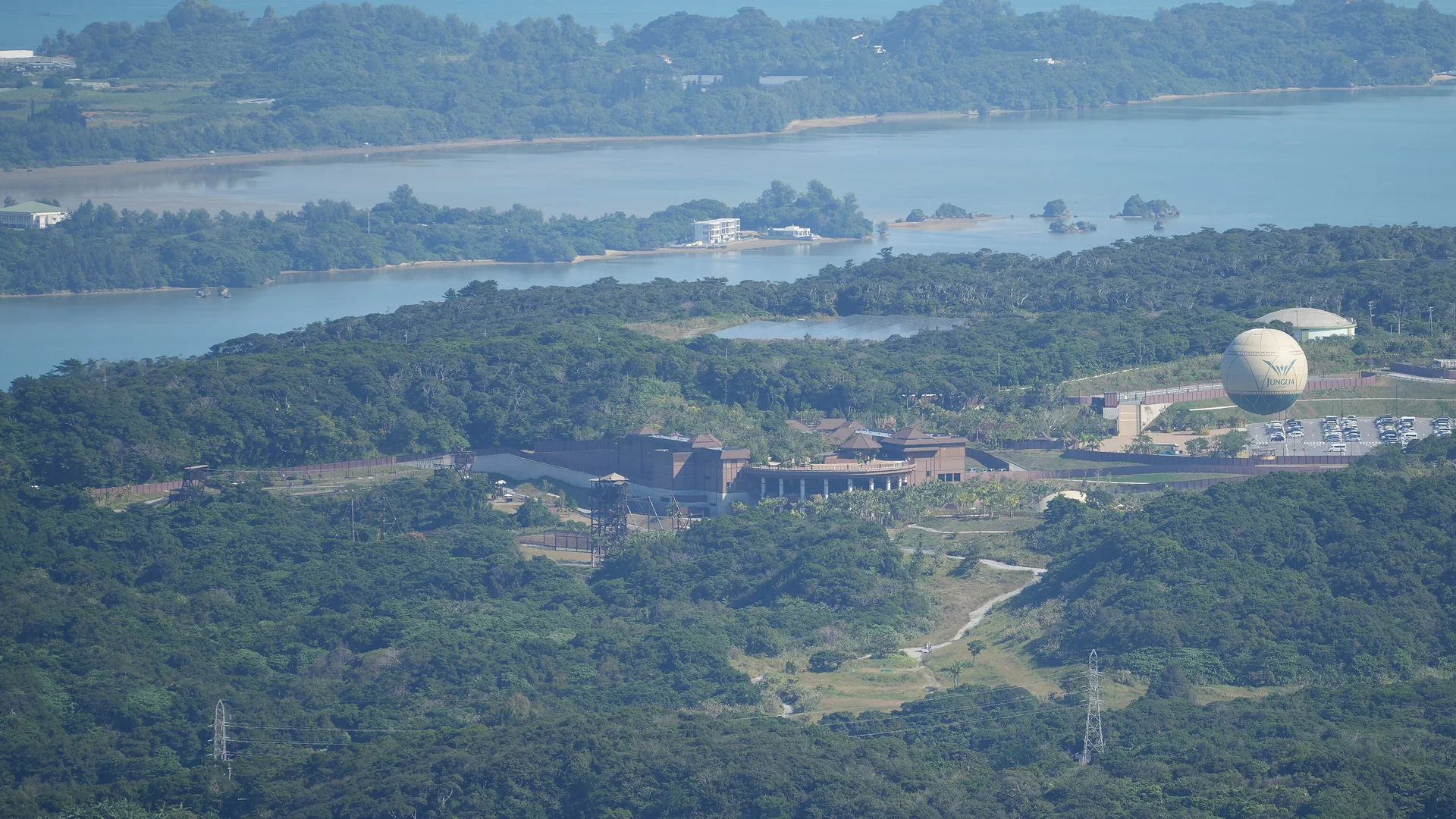 A view from a mountain over the Yanbaru forest of northern Okinawa, with the park's brown entrance buildings and a large pale balloon among the trees above a blue bay with small islands