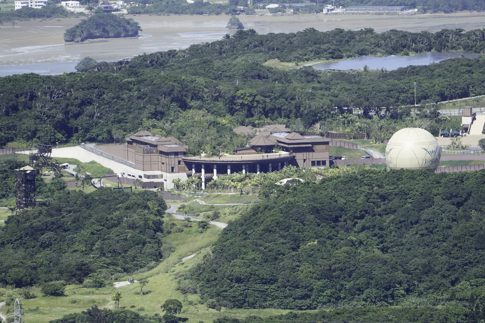 The park's brown entrance buildings and a large pale balloon among dense green forest, with a lake and a tidal bay behind, seen from a hilltop in northern Okinawa